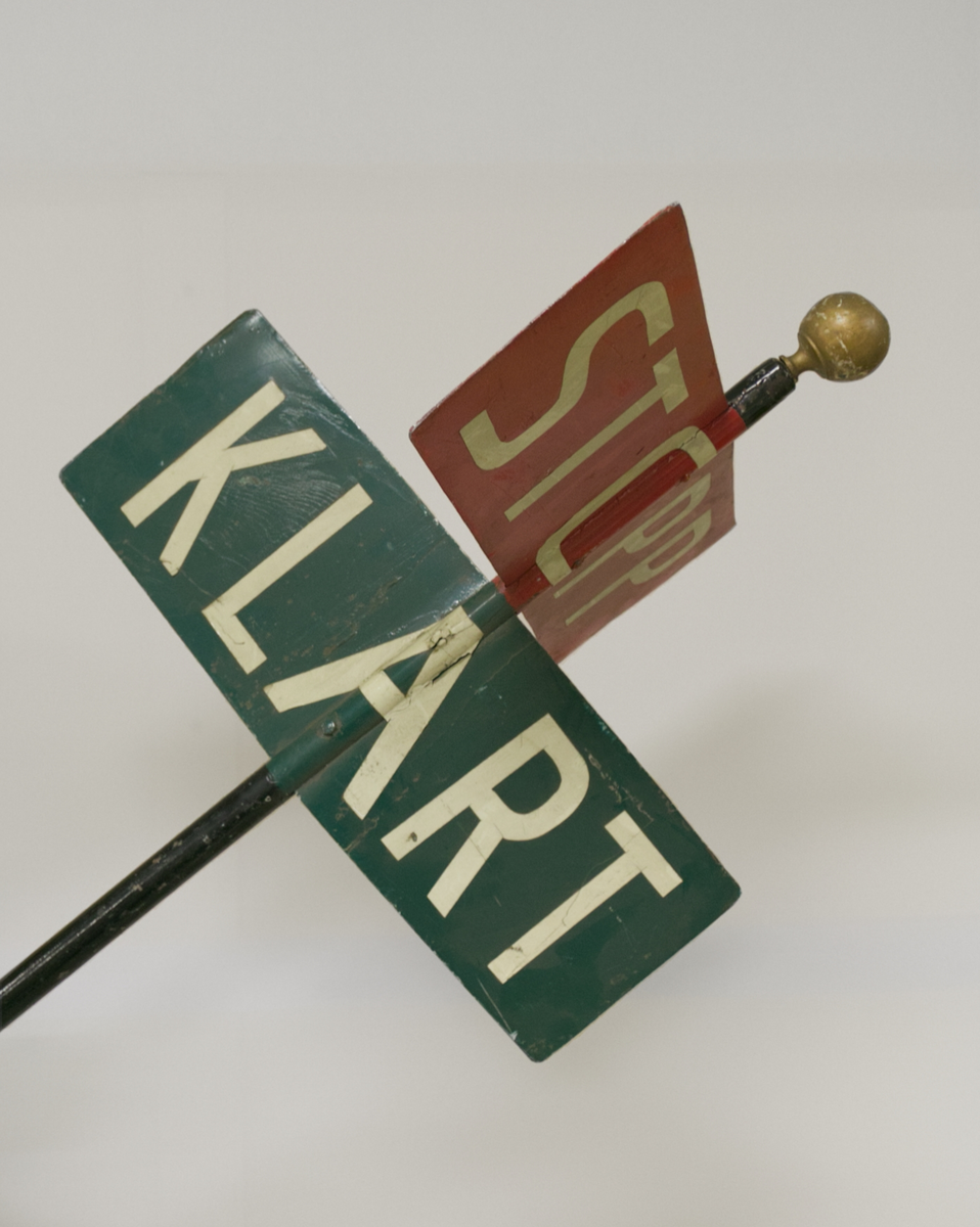 To rektangulære metallskilt. Et rødt skilt med teksten STOPP og et grønt skilt med teksten KLART. Skiltene er montert i hver sin retning på en høy stang med håndtak, slik at skiltene enkelt kan dreies rundt.