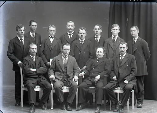 Ateljébild. Beställare: Falkenbergs Typografklubb. Gruppbild av tolv unga och några äldre män i helfigur varav fyra sitter.