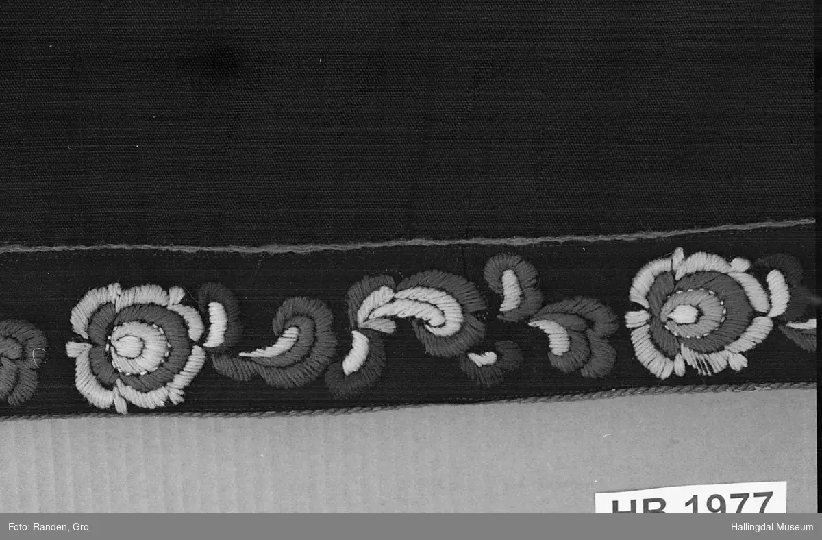 Sort ullstoff. Nederst påsydd bånd med ranke- og rosemønster. Ullgarnsbroderi i plattsøm på sort klede.