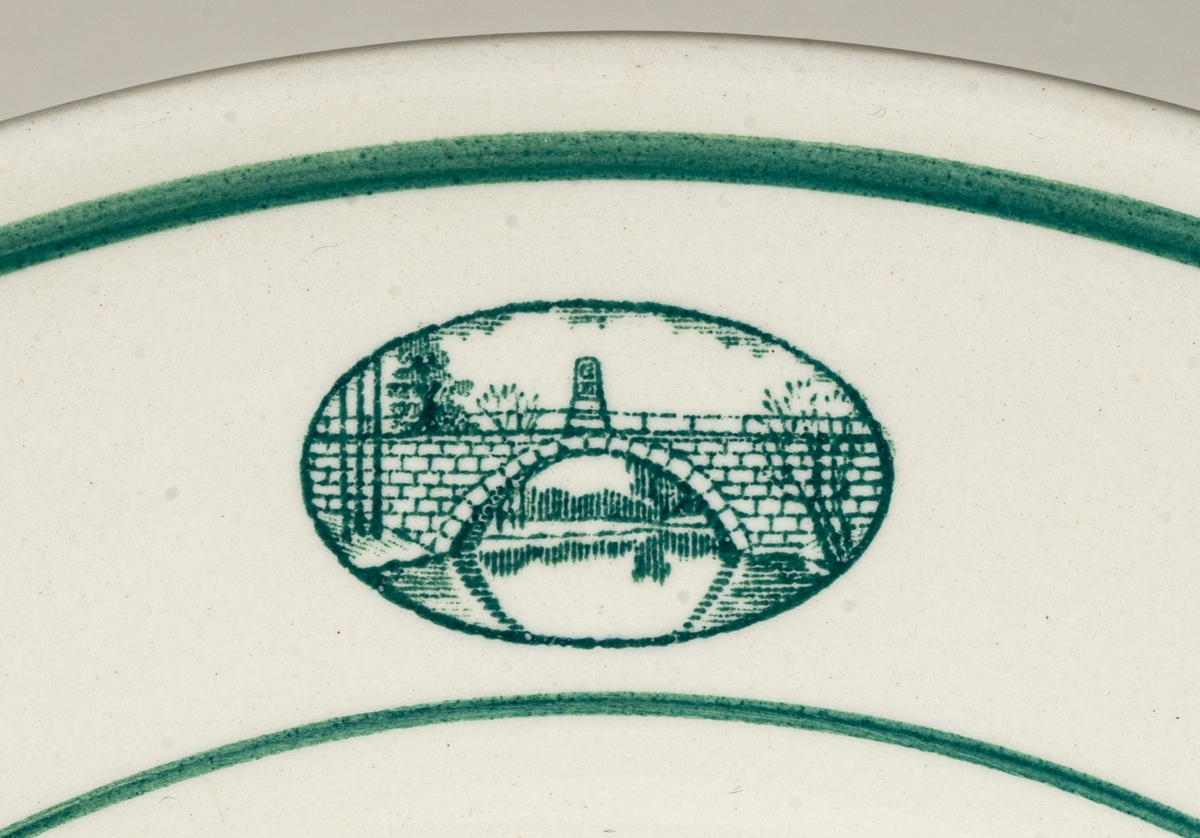 Förrättstallrik till matservis med bild/logotype på brämen föreställande Gustavsbro vid Gävles västra utfart, en bro över Gavleån som byggdes 1782. För vem och varför servisen tillverkades är okänt. Dekoren av två linjer runt brämen samt logotypen är i grönt.