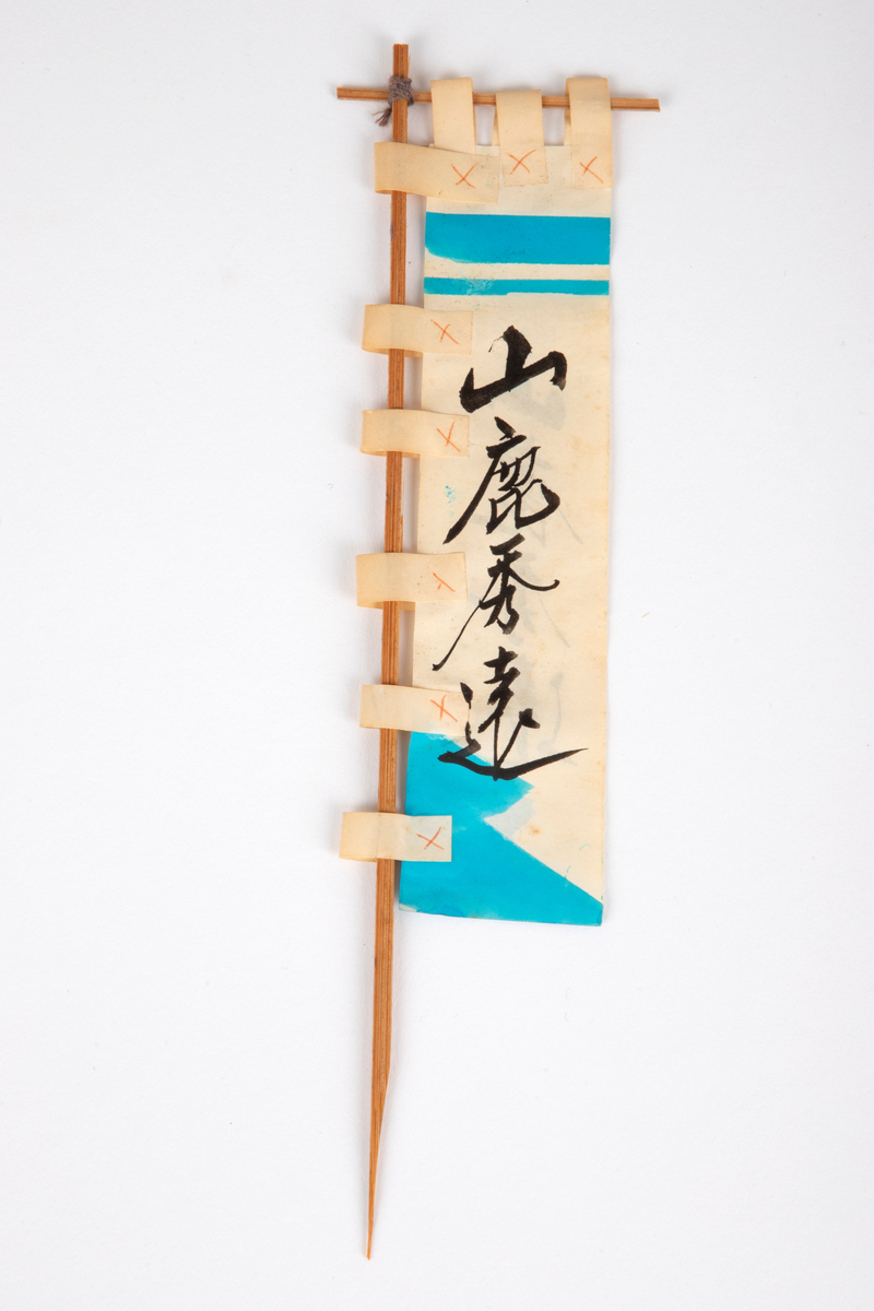 Banner i papir festet til to trepinner. Banneret er hvitt med blå og oransje mønster og svarte japanske tegn. Hører til papirdukken (NK1991-238-001).