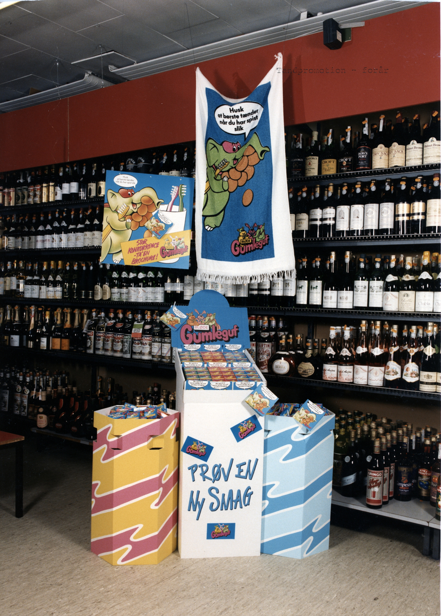 Bild från affär. Gumleguf, tre stycken säljställ står framför hyllor med vinflaskor. Badlakan hänger ovanför.