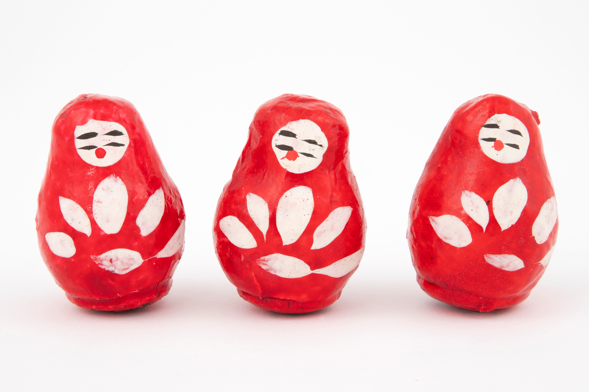 Tre identiske, pæreformede dukker (onna daruma) i pappmasjé. Dukkene er rødmalte med hvit ansikt og hvit bladdekor på fremsiden.