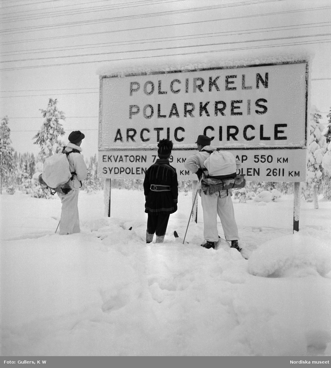 Två vitklädda skidåkare och en man i samisk dräkt vid Polcirkeln som utmärkts med en skylt.
