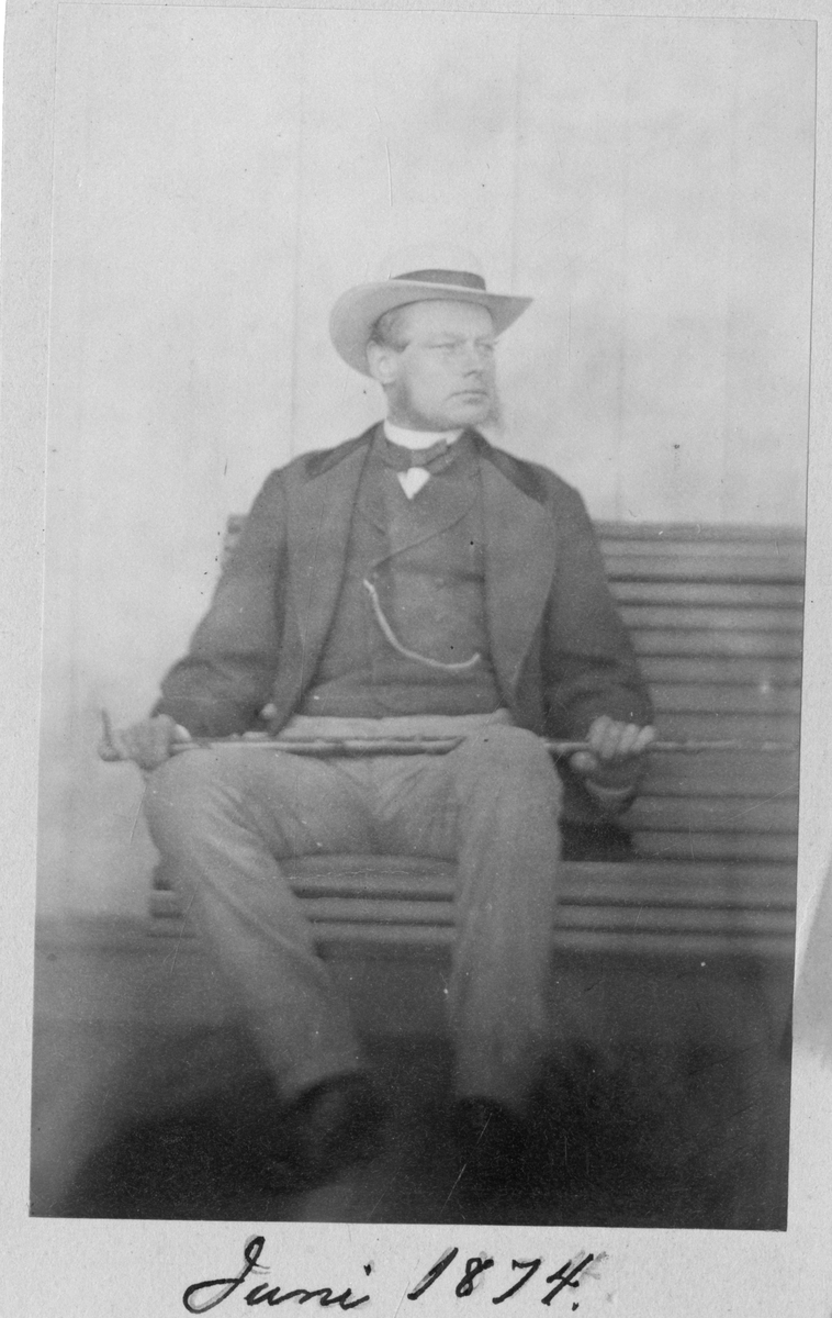 Foto av mann sittende på benk med en spasertokk i hendene. Tatt på Dal gård i 1874