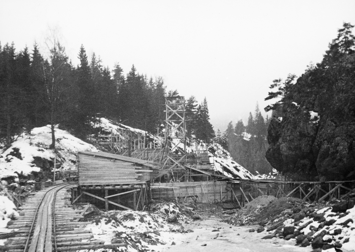 Fra bygginga av en ny dam ved Sølvstufossen, øverst i Ågårdselva i Tune i Østfold, som er en del av Nedre Glommas vestre løp. Anleggsarbeidet startet vinteren 1936, i en periode da det knapt rant vann i elveløpet. Dette fotografiet er tatt i motstrøms retning. Rampa med skinnegang langs nordre elvebredd (til venstre i bildet) ble brukt til å trille materialer og redskap i vogner til og fra anleggsområdet. Inntil denne rampa hadde anleggsarbeiderne reist et bordskur med pulttak. Bakenfor skimter vi forskalinger til dammurer, og over det hele rager noe som må antas å ha vært et heistårn. Også langs den søndre elvebredden (til høyre i bildet) var det reist ei rampe av tre med en gangbane. Ågårdselva er et cirka fem og en halv kilometer langt vassdrag som renner fra den nordvestre enden av Isnesfjorden (Vestvannet) i Nedre Glommas vestre løp, sørvestover gjennem berglendt terreng mot innsjøen Visterflo. Høydeforskjellen mellom Isnesfjorden og Visterflo er på bortimot 25 meter. Ågårdselva har tre fossefall, det øverste her ved Sølvstu, deretter ved Valbrekke og nederst ved Solli. Behovet for den dambygginga vi ser på dette fotografiet var forårsaket av selskapene Borregaards og Hafslunds inngrep i Glommas østre løp, hovedløpet, som i lavvannsperioder gjorde det svært vanskelig å få tømmer som skulle til bedrifter nedenfor Sarpsfossen via Mingevannet, Isnesfjorden og tømmertunnelen til Eidet gjennom det trange sundet ved Trøsken. Dette forsøkte man å løse ved å heve vannspeilet i Isnesfjorden med en dam som skulle plasseres 10-15 meter ovenfor den eksisterende dammen ved Sølvstufossen. Se mer informasjon under fanen «Opplysninger».