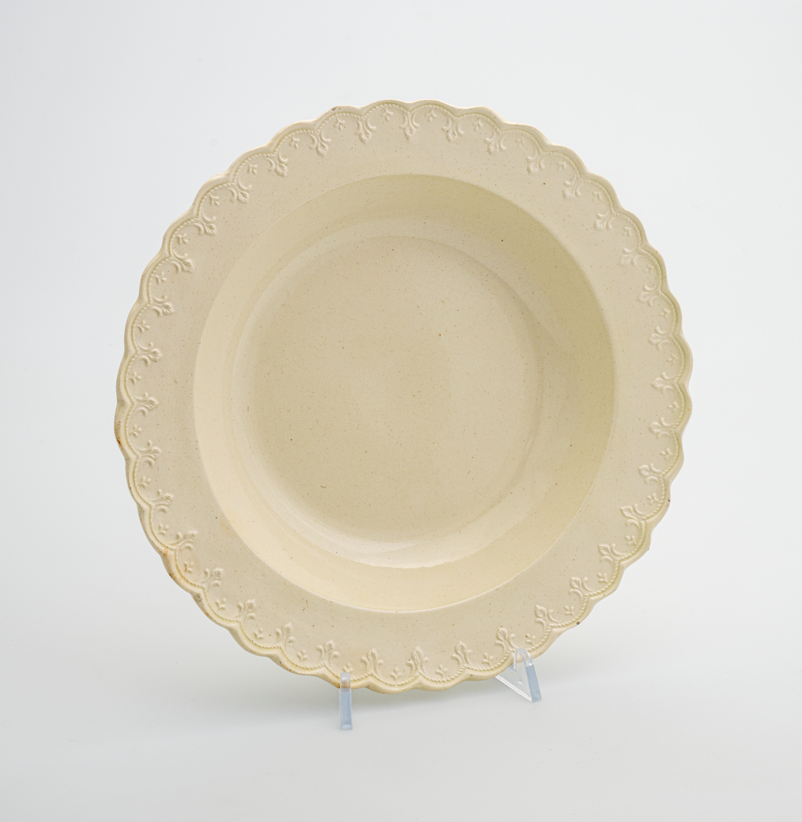 En dyp tallerken av glasert steintøy (creamware). Den er hvitgul på farge (ensfarget). Langs kanten er det tungekant. Innenfor tungekanten er det støpt dekor i form av en hjertebord med stiliserte blomster.