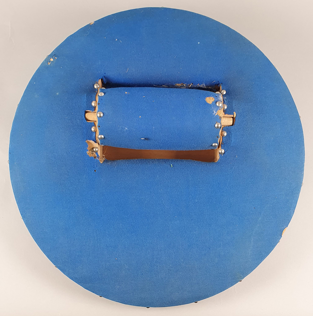 Rundt kniplingsbord av tre trukket med blå filt. En sylinderformet nålepute i midten. Til kniplingsbordet følger et trekk av bomullslerret som er brodert med blå initialer og har blå bånd langs kanten, som kan knytes rundt bordet.