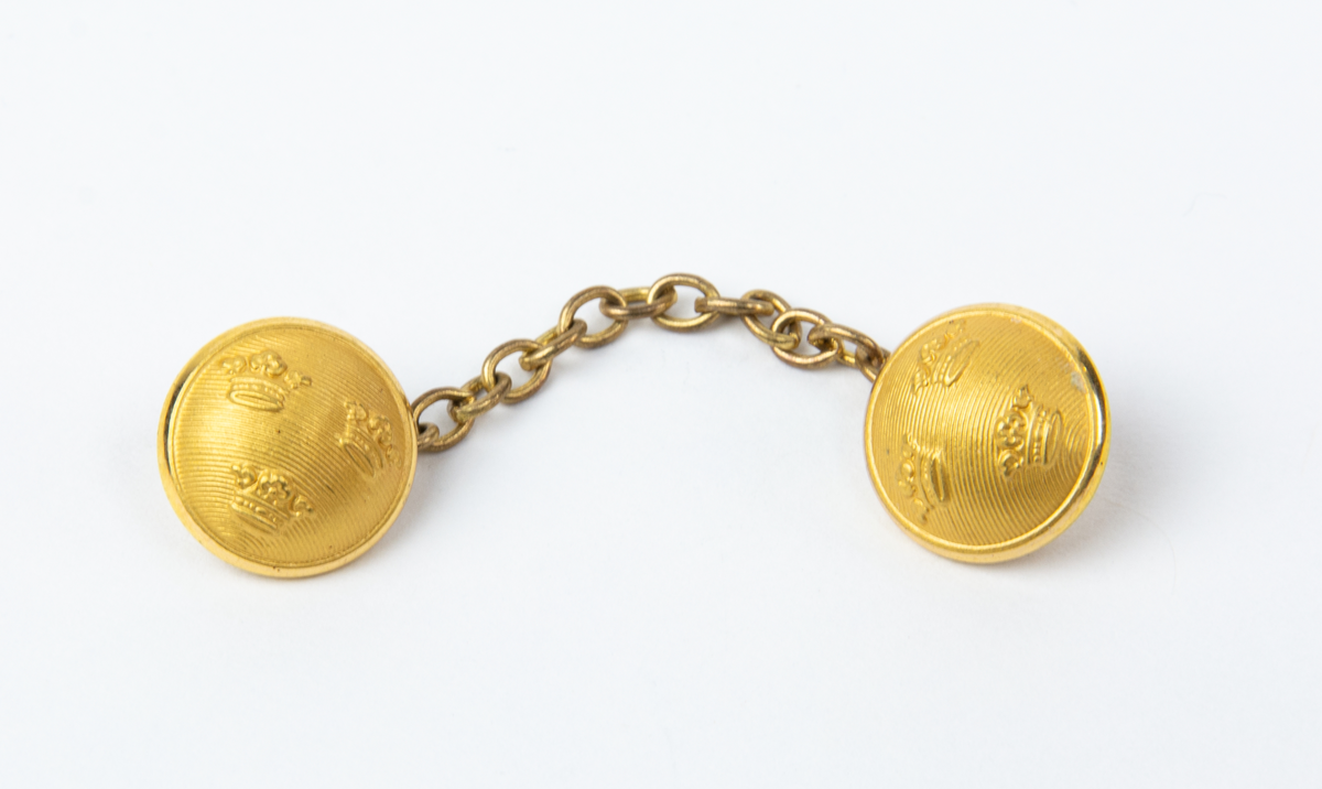 Länkknapp för mässjacka. Två guldfärgade räfflade knappar med tre kronor. Knapparna är sammanlänkade med en kedja. Användes för att hålla samman mässjacka.