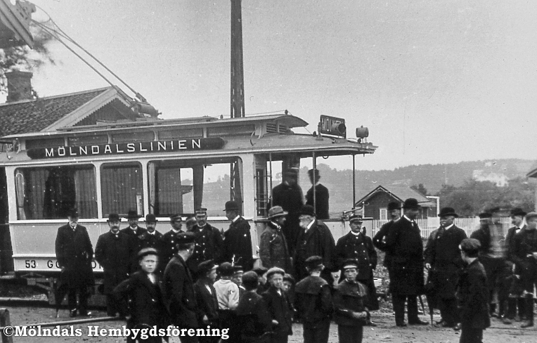 Mölndalsbro i Mölndal, år 1907. Spårvägslinjen invigs. Intill en spårvagn har en samling herrar och gossar samlats.