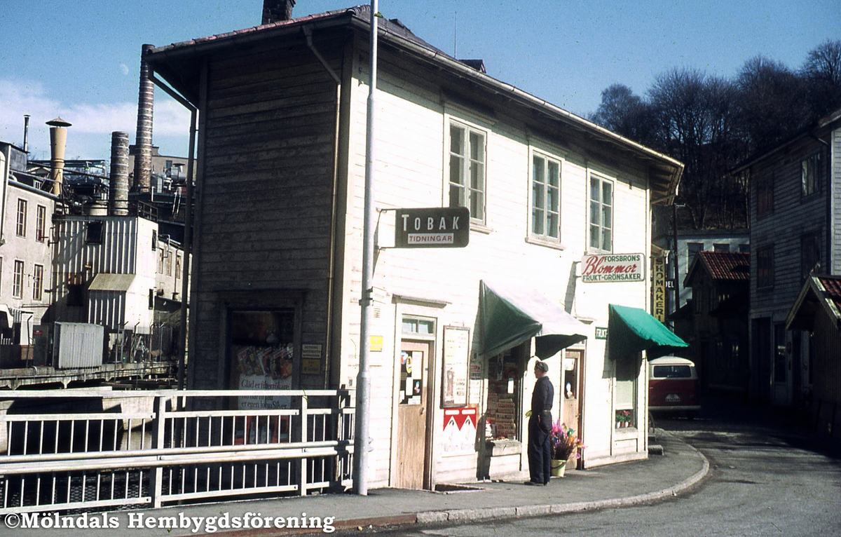 Tobaksaffär och blomsteraffär i huset Götaforsliden 1-5 (nuvarande Kråkans Krog) i Mölndals Kvarnby, 1970-tal. Vy från Forsebron. Till vänster ses industribyggnad tillhörande SOAB. Kv 45:14.