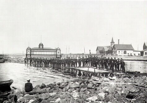 En stor samling män i uniform, kanske militärer eller sjömän, står på en brygga ut mot det lilla skäret vid Barnens strad i Varberg. Nere till vänster sitter en man i skärmmössa vid en eka och betraktar dem. I bakgrunden ses till höger varmbadhuset från 1866 och till vänster kallbadhuset som fanns mellan 1886-1902.