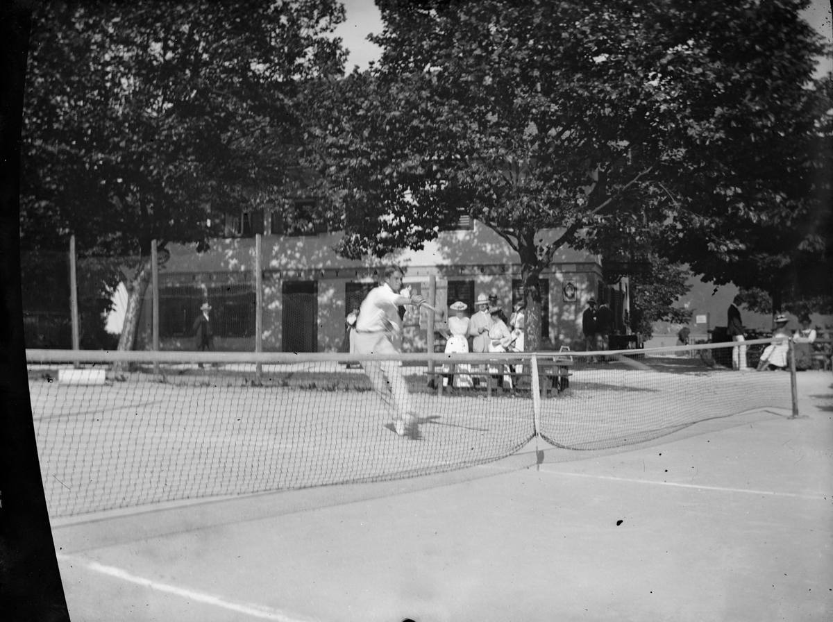 En man spelar tennis på en bana i en park i Wien, 1910-tal. I bakgrunden åskådare och en restaurang.