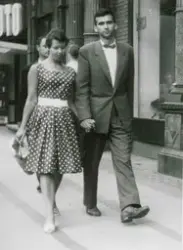 Et pent kledt ungt par spaserer hånd i hånd i en gate. Svein