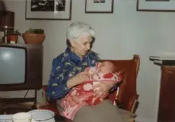 En eldre kvinne holder et spedbarn. I bakgrunnen et fjernsyn