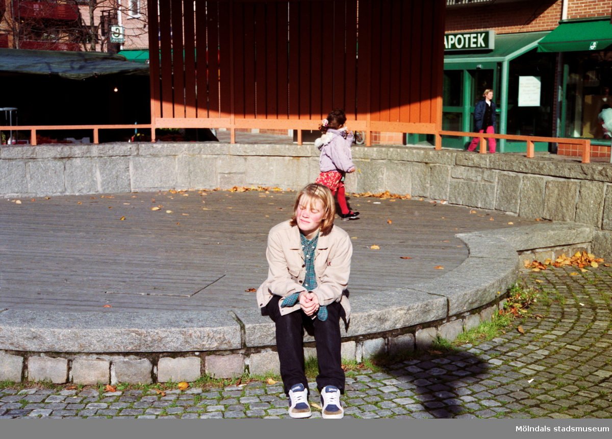 En tjej sitter på en stenmur vid Alberts torgs scen. Mölndalsbro i dag - ett skolpedagogiskt dokumentationsprojekt på Mölndals museum under oktober 1996. 1996_0991-1004 är gjorda av högstadieelever från Kvarnbyskolan 9A, grupp 5. Se även 1996_0913-0940, gruppbilder på klasserna 1996_1382-1405 samt bilder från den färdiga utställningen 1996_1358-1381.