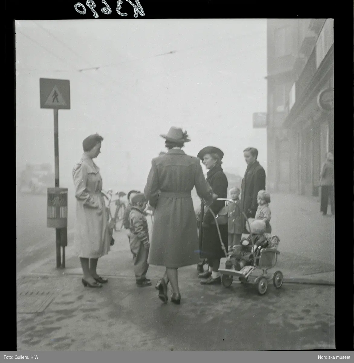 542/B Dimma. Några kvinnor med barn och barnvagnar har stannat för att prata på trottoaren.