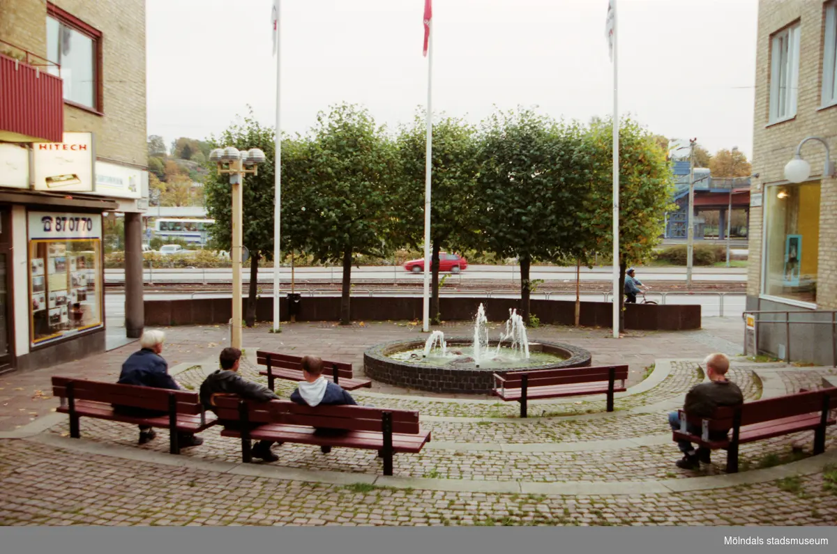 Personer sitter på bänkar i början av Brogatan. Göteborgsvägen passerar i bakgrunden. Mölndalsbro i dag - ett skolpedagogiskt dokumentationsprojekt på Mölndals museum under oktober 1996. 1996_1246-1263 är gjorda av högstadieelever från Åbyskolan 8A, grupp 1. Se även 1996_0913-0940.