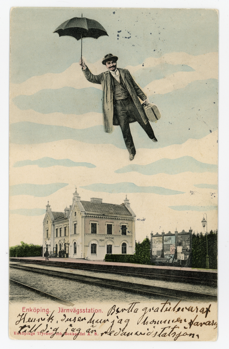 Vykort med motiv av Järnvägsstatione, Enköping. Vykortet är handkolorerat och uppe på himmelen flyger en man med väska & paraply som är ditretuscherad i efterhand. Vykortet är poststämplat 1900, datumet är oläsligt.