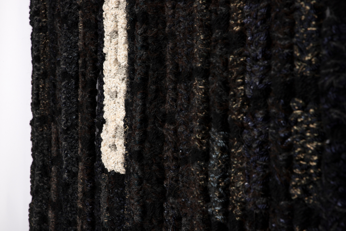 Billedvev i sort bestående av fritthengende remser av ullgarn og tråder i metall og silke. Med omtrent ti centimeters mellomrom er båndene snevret inn slik at det oppstår en rytme i flatene. På de tre miderste remsene er det smettet inn et felt i hvitt.