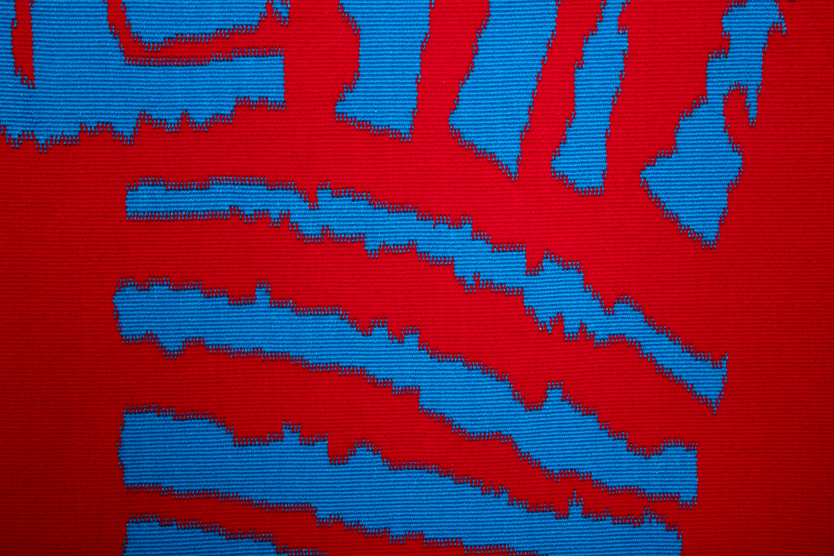 Rektangulær billedved i rødt og blått. Portrettet viser ansikt og hals bygget opp av rette og vinklede former i blå. Formene har ujevne kanter. Bakgrunnen er rød, inrammet av et blått felt. Figuren er plasert i øvre høyre hjørne. Bunnen er i ull, og mønster i silke.