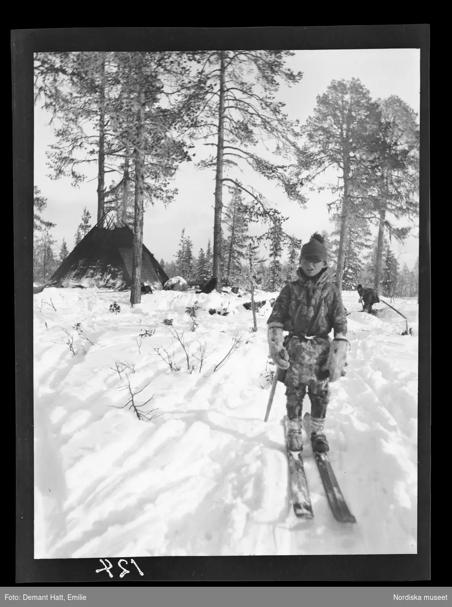 Pojke, Andaras (Nilsa), på skidor i snön, troligen under höstflyttningen vid Laimo. I bakgrunden ses en kåta och en person som gräver snö (till kaffekokning). Bilden ingår i en serie fotografier tagna av Emilie Demant Hatt i Sapmi mellan åren 1907 och 1916.