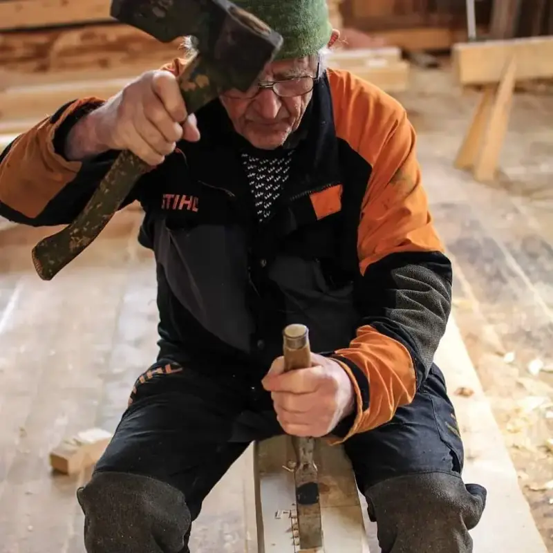 Håndverker Jon Bojer Godal sitter tvers over på et materiale. Han har en øks i den ene hånden og en stemjern i den andre. Han skal til å slå stemjernet med den butte enden av øksen.