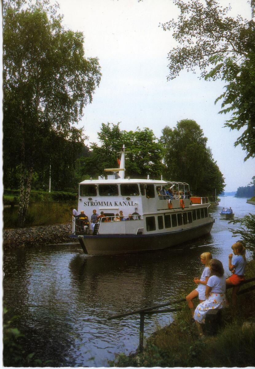 Passagerarbåten Strömma Kanal passerar Strömma kanal i västlig riktning. I förgrunden t h står Sigvard Lönnqvist. Dalholmen skymtar i bakgrunden till höger vid kanalens östra utlopp i Brevikssundet.