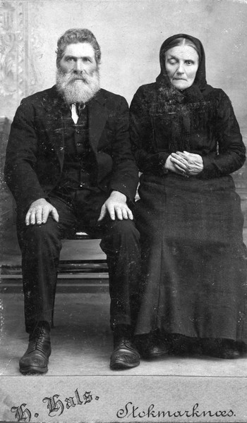Helportrett av eldre ukjent mann og kvinne sittende på benk i fotostudio. Fotograf H.Hals. Stokmarknes. Trolig Stefanus Steffensen, Breivik.