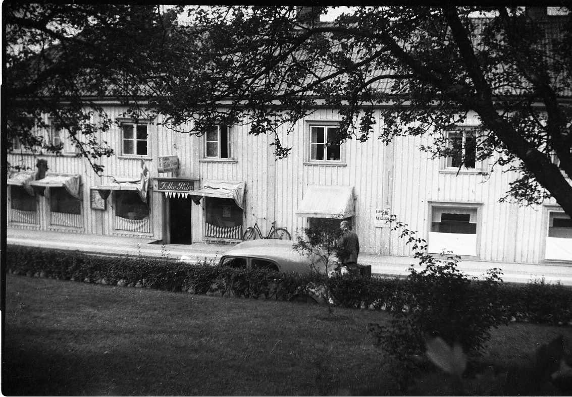 En fastighet på Brahegatan i Gränna, fotograferat mellan träd på motsatta sidan. Skylt för Folke Palms garn- och manufakturaffär.