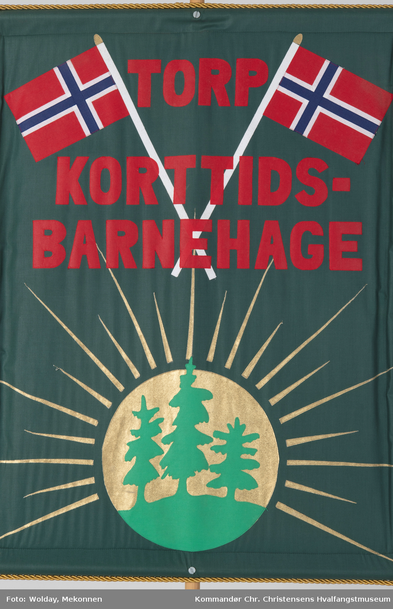 Mørkegrønn bakgrunn med rød skrift. Dekortert med norske flagg og en sol med grantrær. Festet til en rundstokk.