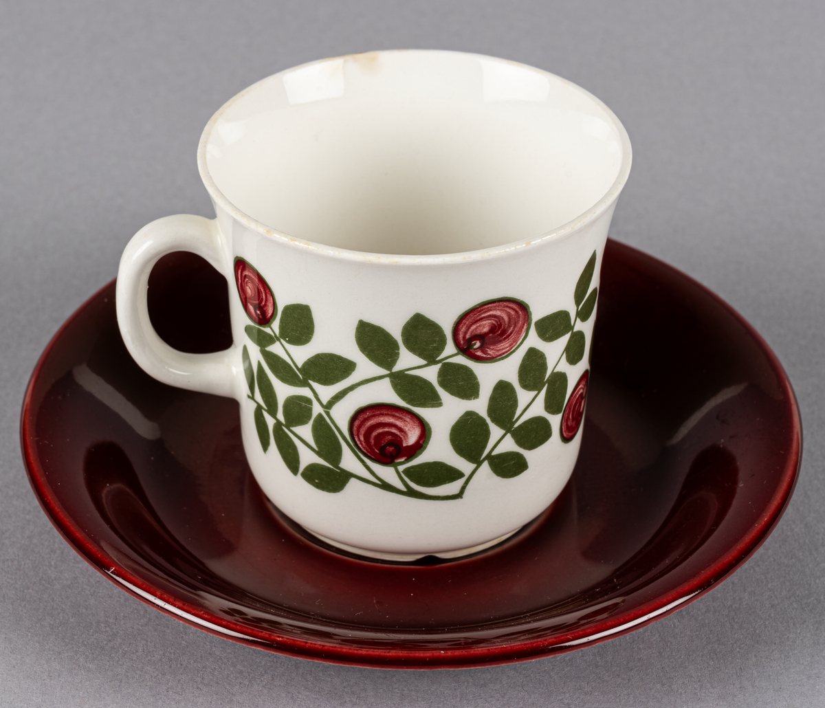 Kaffekopp med fat, i flintgods. Modell EF, dekor Klarbär. Vit glasyr med röda bär och grönt bladmönster i grönt, fatet mörkrött. Otydlig stämpel på fatets undersida. Tillverkad troligtvis på 1960-talet.