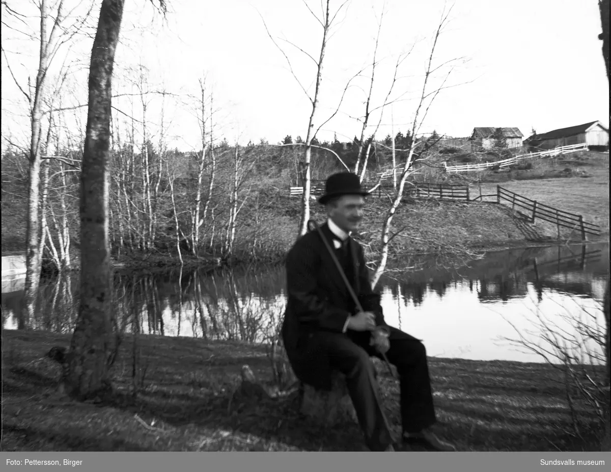 En man med hatt, käpp och helgdagskläder sitter på en stubbe i lantlig omgivning. Bakom syns ett stilla vattendrag, inhägnader i trä och enklare byggnader, kanske en fäbodvall.