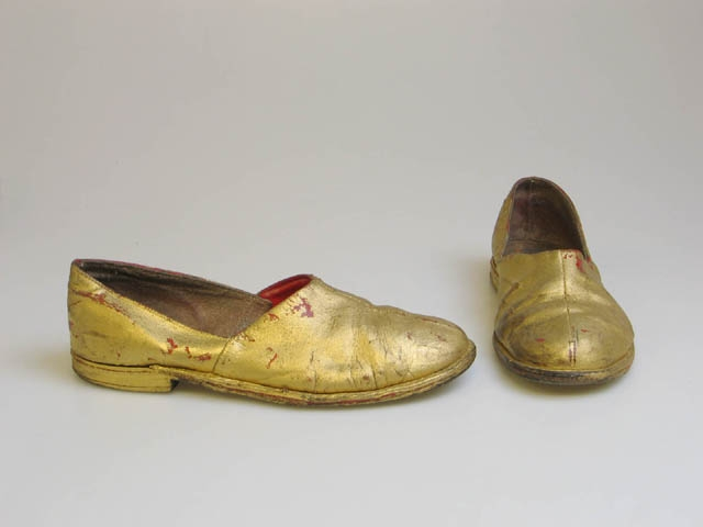 Guldfärgade skor. Tillhör maskeraddräkt LAM024974.
Ett par guldfärgade skor. Mått: L 27, B 9,5, Storlek 39. 
Förvärvades i samband med utställningen "Maskerad" på Landskrona museum 21/11 2004-23/1 2005.
Från början använd på en maskerad i Landskrona på 1950-talet.
Bilder från maskeraden där kläderna användes finns i museets bilddatabas, B 1005128-1005130.