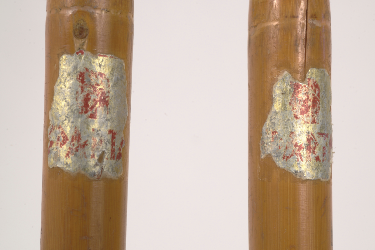 En av to skistaver, den andre A.06019-03.a. Selve staven er brun bambus. Håndtak, reimer og feste til trinse hvitt skinn. Metallpigg på enden. Trinsen av tre.
