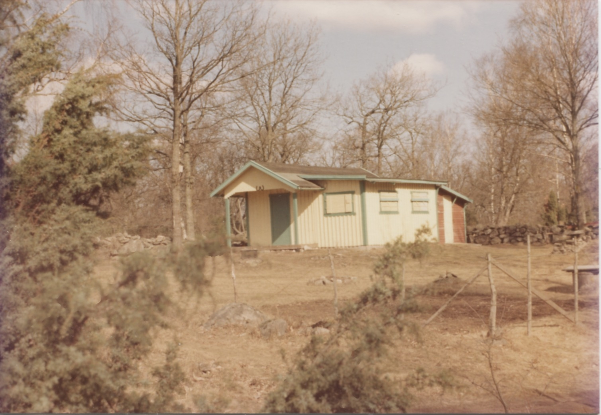 En gul/grön sommarstuga med stängda fönsterluckor och staket runt tomten, 1960-tal. Stugan tillhörde Högen och låg där nuvarande Livereds kulturstig går.