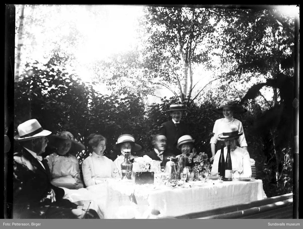Gruppbild med glada män och kvinnor vid ett dukat bord i trädgården.