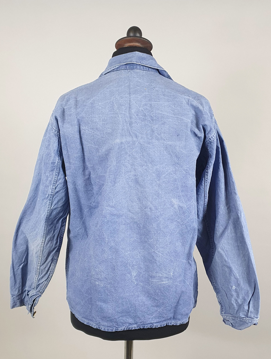 Skjorte (busserull) av blå dongeri. Åpning foran med knappestolep og skjortekrage. Trykknapper av metall foran og på mansjettene.