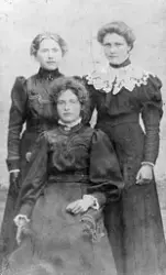 Portrett av tre piker, en sitter, de har høyhalsede kjoler. 