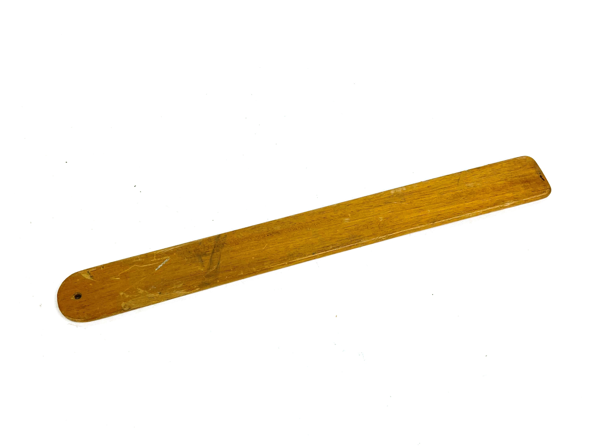 Packsticka i trä, avlång med rundad form i ena änden. Ett mindre hål i den rundade formen.