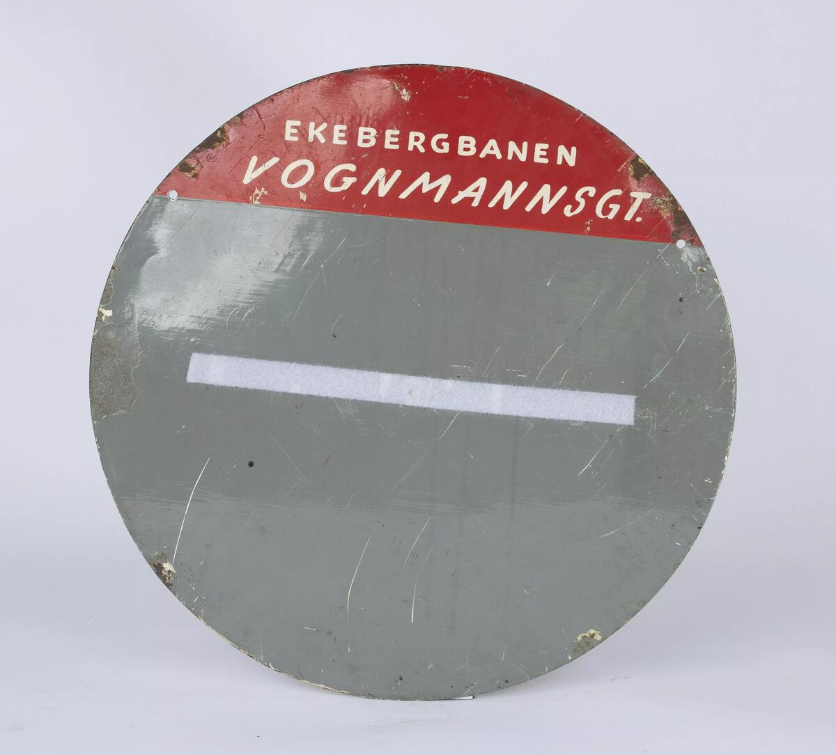 Rundt linjeskilt for Ekebergbanen plassert i vognmannsgata og viser tider for linje til Ljabru. Skiltet er av stål og er malt i en kremhvit og rød farge.