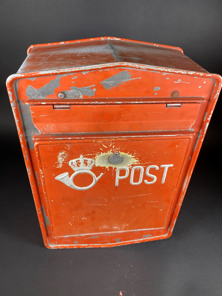 Rød postkasse med innstikksluke, merket med Postens logo og "POST". Postkassen har slitasjemerker etter bruk og noe malingsflass. Anheng for montering på baksiden.