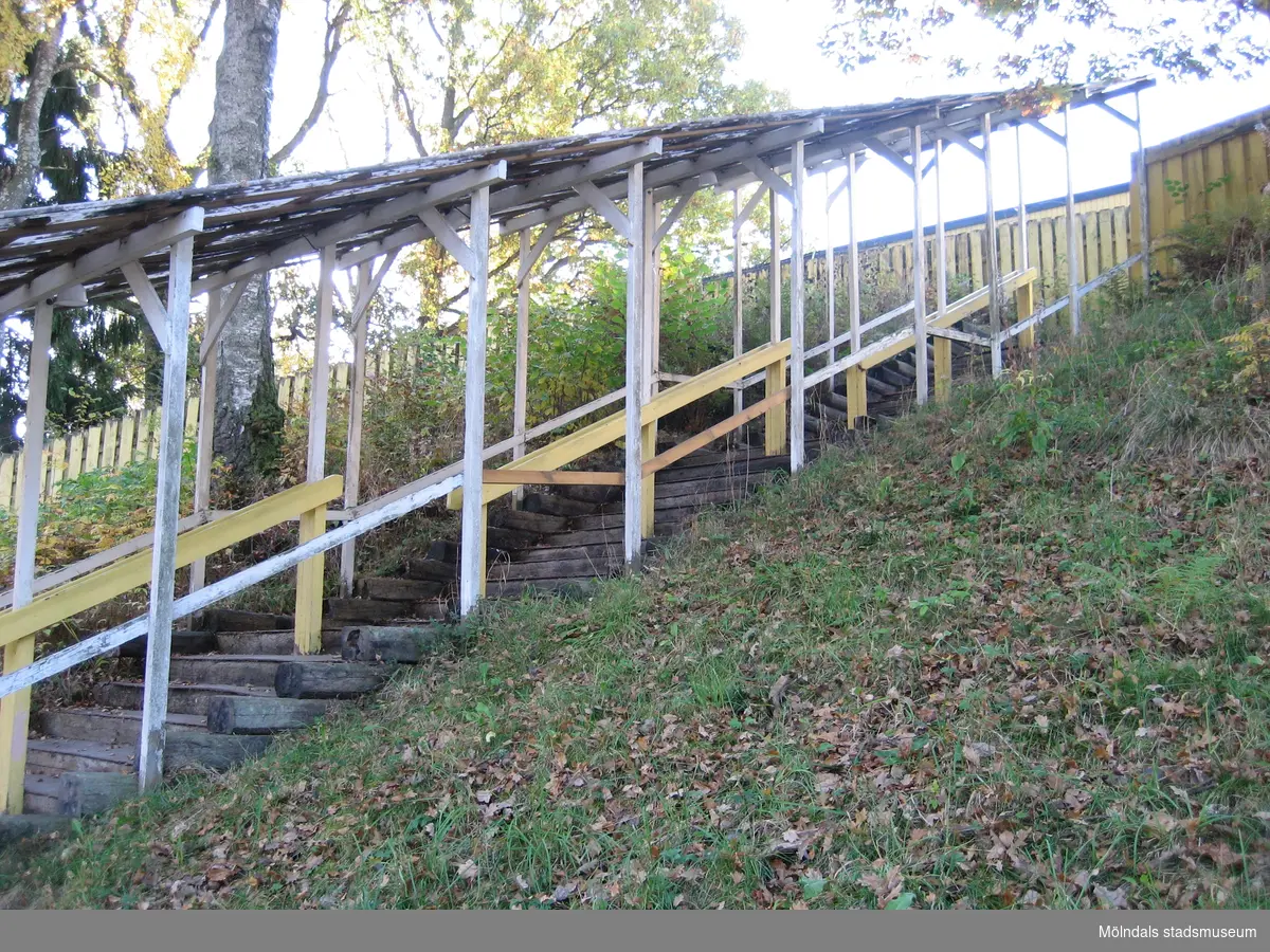 En sluttande fristående trappa med tak, sedd från höger, där människor kan köa till alternativt lämna Hällesåkers dansbana år 2010. Området är omgivet av växtlighet samt är inhägnat med staket.
Relaterade motiv: 2024_1119 - 1137.