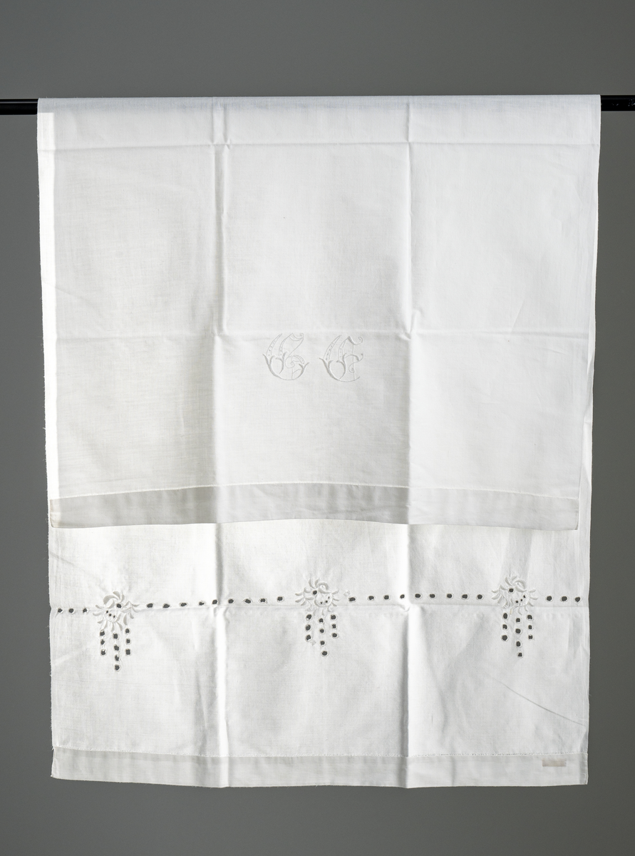 Pyntehåndkle i hvit bomull med monogram "G.E." i nedre kant og engelskbroderi m. hullsøm i den andre enden. Dobbel kant i begge kortender. Vevd i hel bredde.
