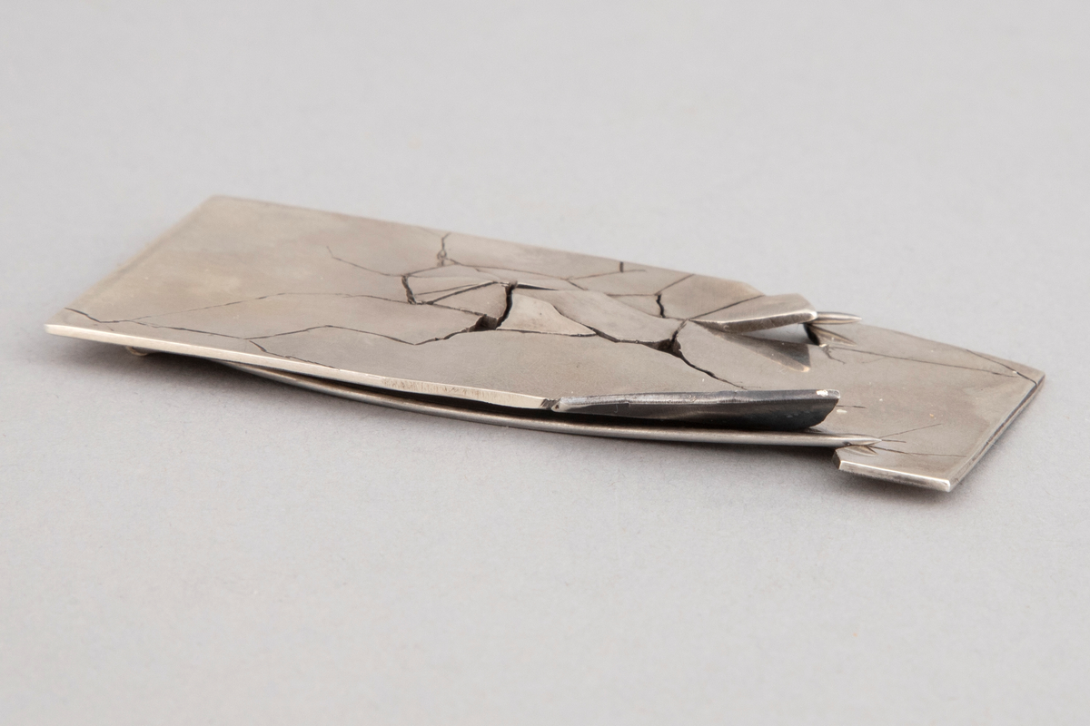 Rektangulær brosje i sterling sølv med sprekker og brudd på framsiden. Dobbel nål på baksiden.