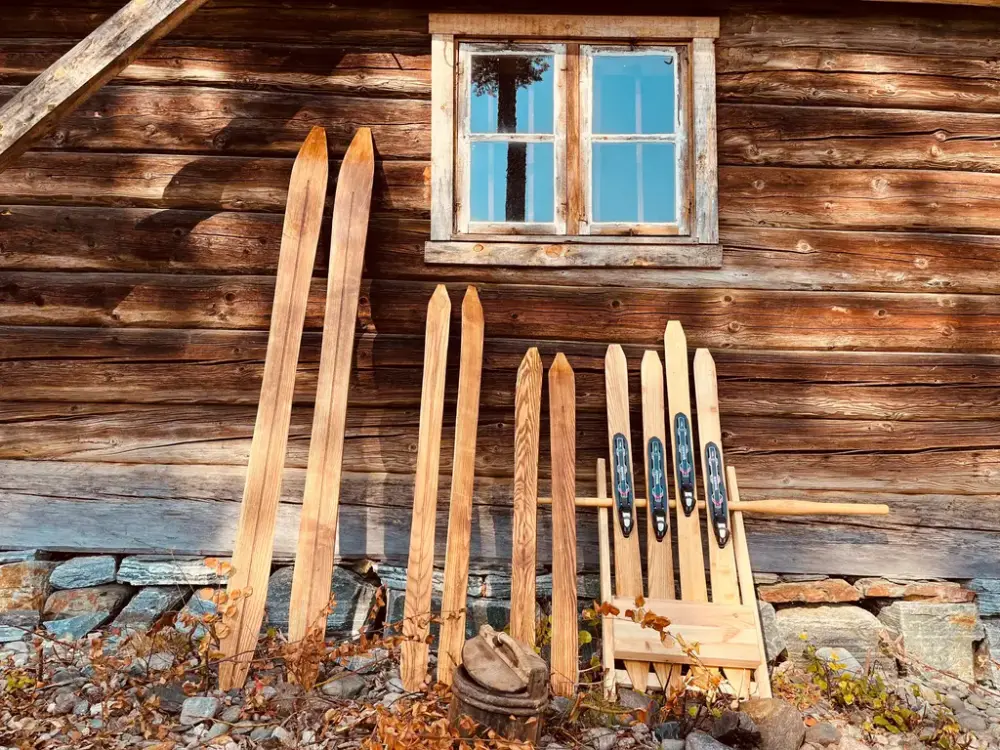 Høst på Rindal skimuseum. 5 par gamle ski står oppstilt mot en husvegg i høstsol.