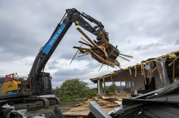 En gul og blå beltemaskin har grabben full av planker som spriker i alle retninger, et delvis revet hus står i bakgrunnen.