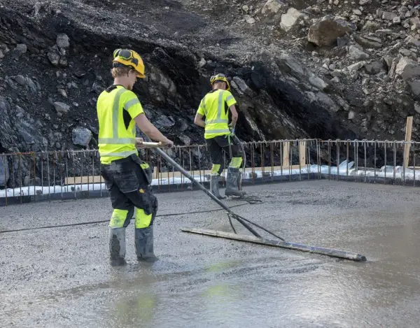 To menn med signalklær og hjelm glatter en stor, nystøpt betongflate.