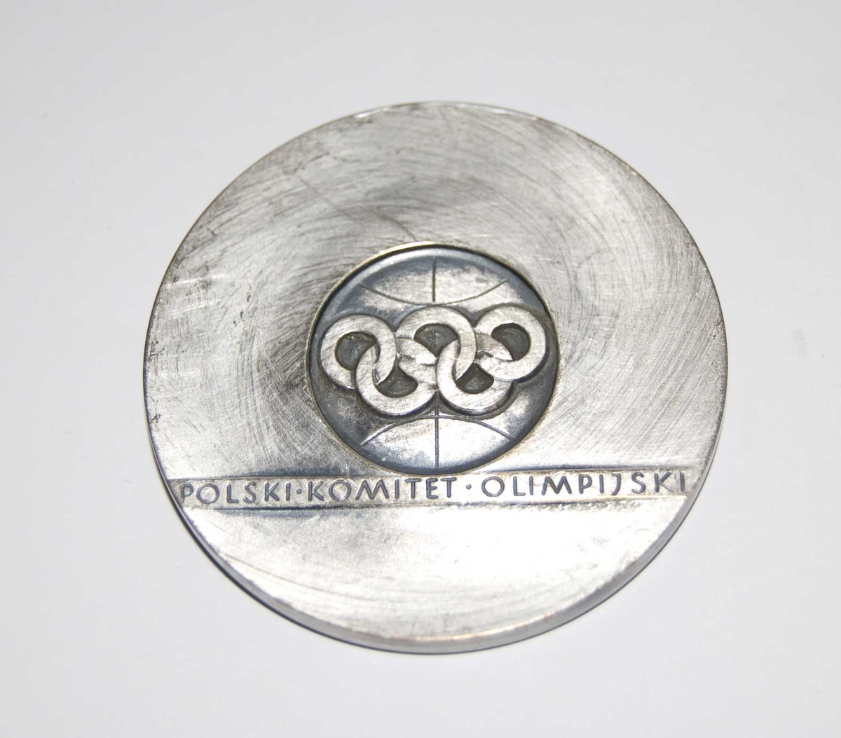 Sølvfarget medalje med motiv av de olympiske ringer og løpende atleter. Det er også et stilisert mønster/motiv på medaljen. Det følger med en rød eske til medaljen.