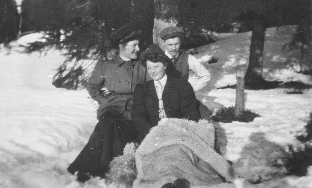 To kvinner og en mann sitter i snøen under en gran. Påsken i Nittedal.